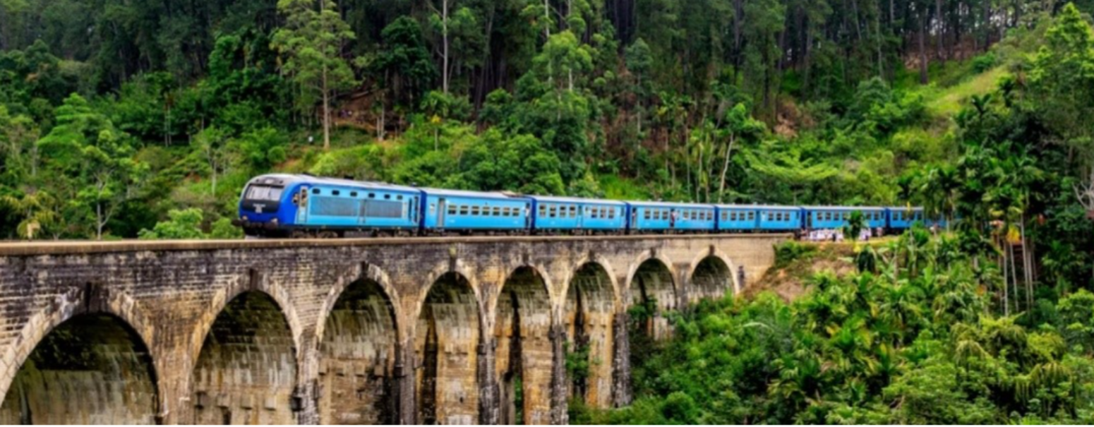 Blue passenger train crossing a stone arched viaduct surrounded by lush green forest and hills.