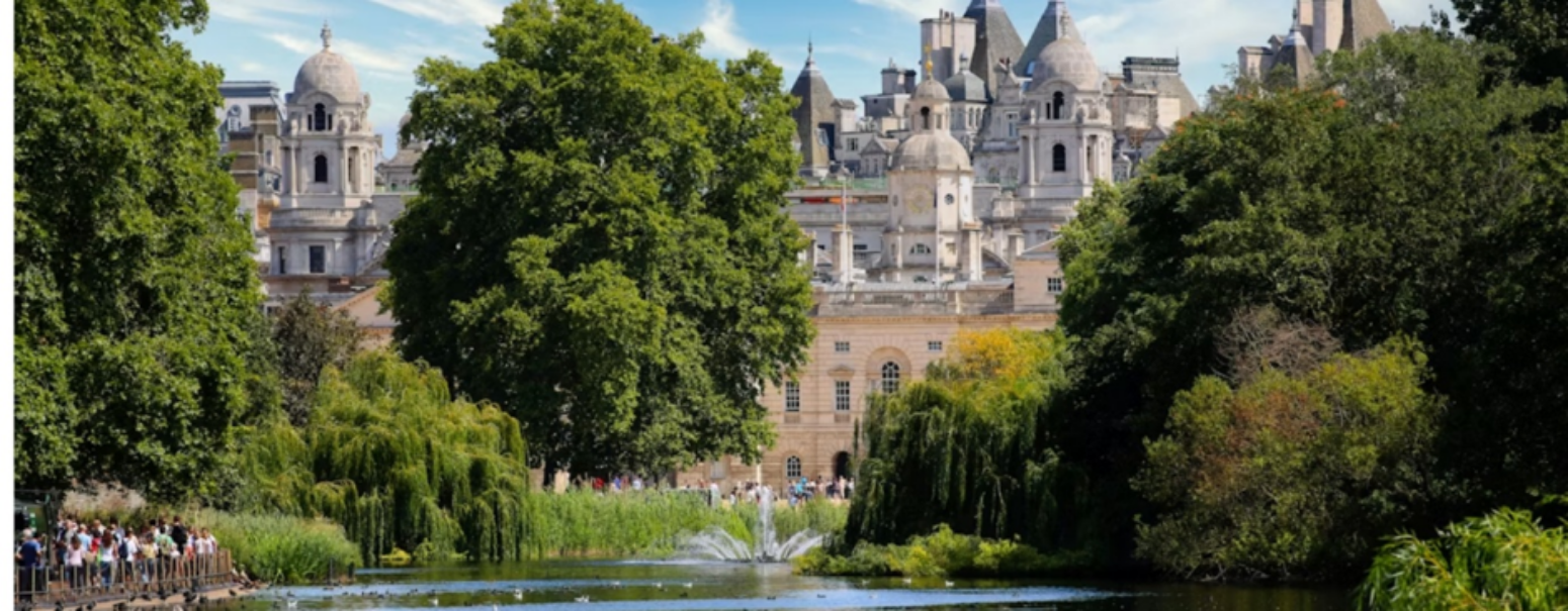 Alt text: "Sunny view across the lake in St James's Park, London, with a fountain spraying water, willow trees along the banks, and the domed towers of Horse Guards and Whitehall buildings visible through the trees in the background