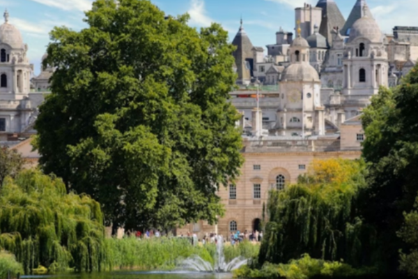 Alt text: "Sunny view across the lake in St James's Park, London, with a fountain spraying water, willow trees along the banks, and the domed towers of Horse Guards and Whitehall buildings visible through the trees in the background