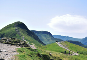 Walkers follow a ridge path along Cat Bells in the Lake District, with layered fells stretching into the distance