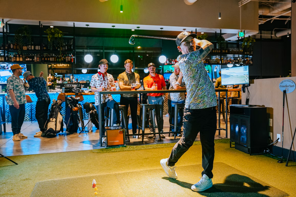 A golfer swings a club indoors while several people watch from a bar area.