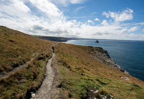A hiker walks a coastal path along grassy cliffs overlooking the sea on the South West Coast Path.