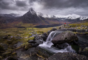 A hiker pauses beside a waterfall beneath the jagged peak of Tryfan in Eryri (Snowdonia).